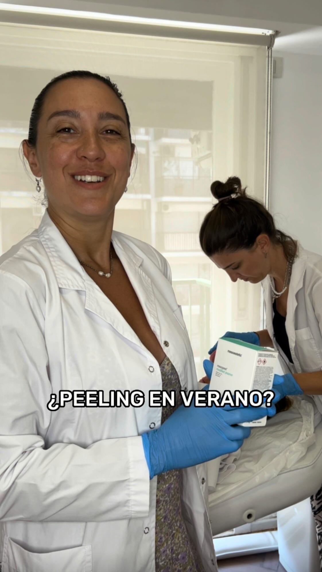 La Dra. Renata Schvartz sonríe con guantes y un producto de peeling mientras una colega prepara a una paciente en la camilla, junto a la ventana
